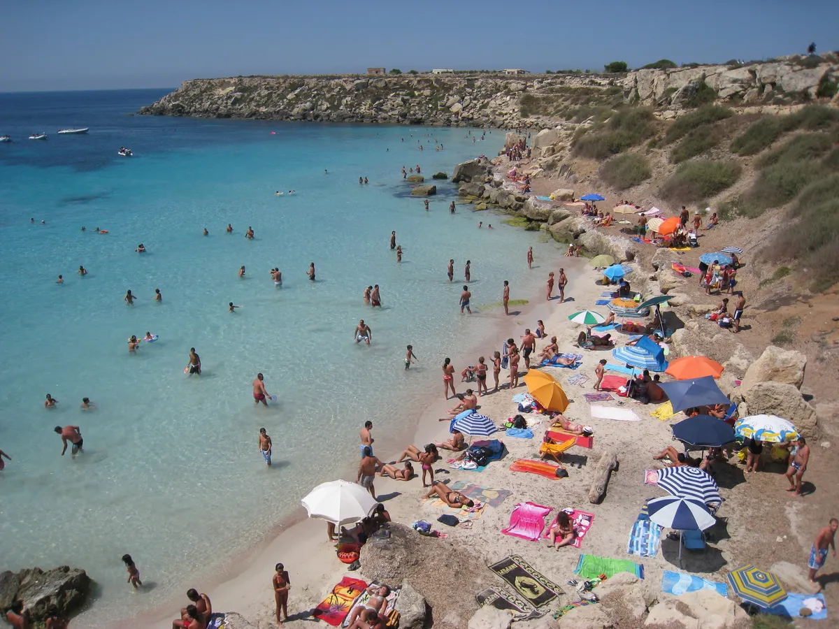 Il piccolo arenile sabbioso di Cala Azzurra a Favignana, con ombrelloni, bagnanti e rocce ai lati della riva.