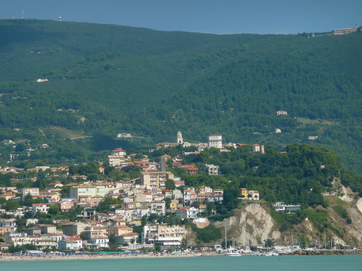 Veduta di Numana con le case sopra la falesia, una spiaggia e il porto turistico in primo piano.