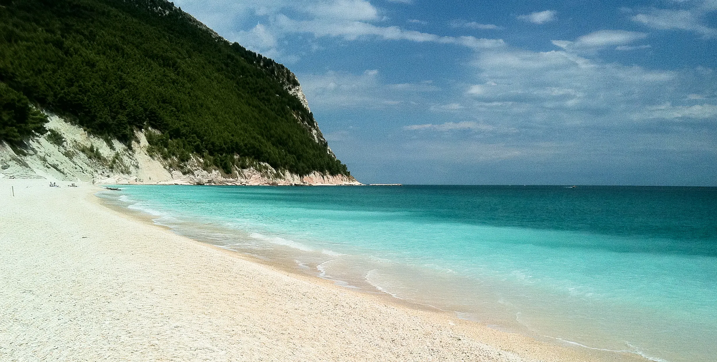 Una spiaggia di Sirolo con ghiaia chiara, mare turchese e il versante alberato del Conero.