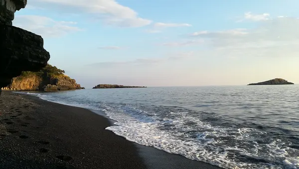 La Spiaggia Nera di Maratea nel giugno 2019, con arenile scuro, parete rocciosa e isolotti sullo sfondo.